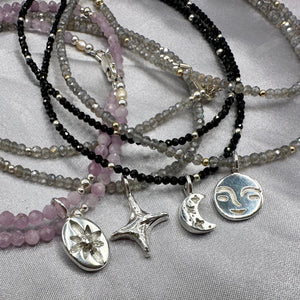 Stack of beaded necklaces with various silver pendants on a light gray fabric background